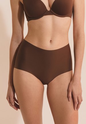 Mujer con ropa interior marrón de cintura alta sin costuras y sujetador a juego, de pie sobre un fondo beige.