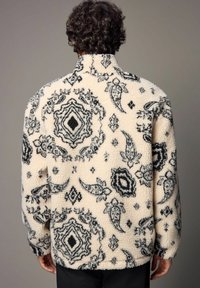 Kremowa polarowa kurtka z czarnymi wzorami w paisley i romby; posiada wysoki kołnierz oraz miękką, teksturowaną powierzchnię.