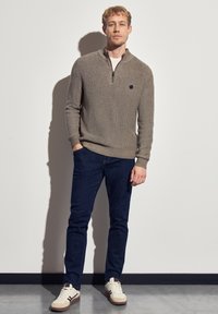 Un sweat à capuche gris en tricot avec une texture en côtes, associé à un jean bleu marine et des baskets beiges. Présente un emblème noir sur la poitrine.