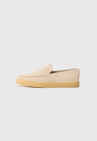 Zapato slip-on de ante beige con punta redondeada y suela de goma marrón claro, mostrado en perfil lateral sobre un fondo blanco.