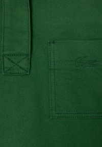 Grüner Stoff mit glatter Textur verfügt über eine genähte Tasche und ein Logo. Der Riemen hat ein X-Stich-Design zur Verstärkung.