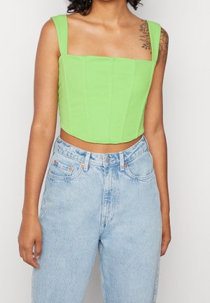 Vrouw die een limoengroen mouwloos cropped top en lichtblauwe high-waisted jeans draagt, met een bloemen tatoeage op haar bovenarm.