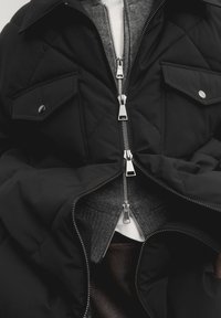 Zwarte gewatteerde jas met zilveren ritsen, drukknoopzakken en een grijze binnenlaag. Heeft een soepele textuur en een gestructureerd ontwerp.