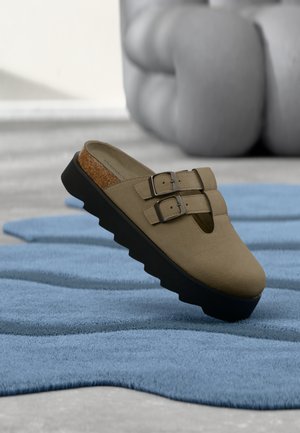 Chaussure en daim marron à enfiler avec deux brides à boucle, un accent en liège et une semelle épaisse en caoutchouc noir, posée sur un tapis texturé bleu.