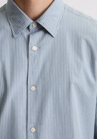 Chemise boutonnée bleu clair avec des rayures verticales blanches, dotée d'un col classique et de deux petits boutons ronds à l'avant. Texture douce.
