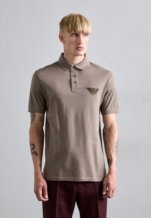 Brun polo skjorta med krage, tre knappar fram och en broderad logotyp på bröstet. Korta ärmar och mjuk bomullstextur.
