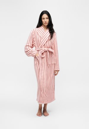 Vrouw met lang donker haar, gekleed in een roze badjas met verticale rode strepen, gestrikt in de taille, blootsvoets staand op een witte achtergrond.