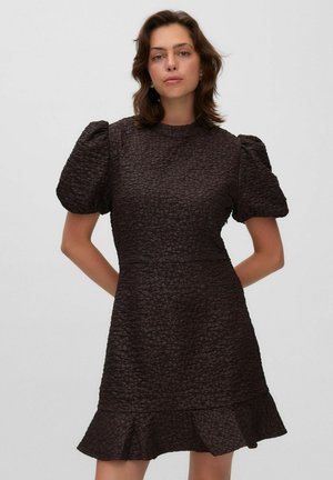 Mujer con cabello castaño lleva un vestido corto oscuro con textura, mangas abullonadas y dobladillo acampanado, de pie con las manos detrás de la espalda.