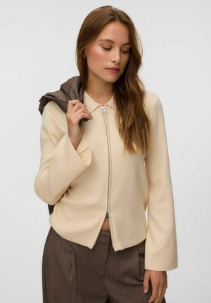 Femme aux longs cheveux bruns porte une veste crème zippée et un pantalon marron, tenant un pull marron sur son épaule devant un fond uni.