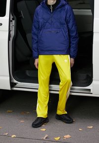 Marinblå puffjacka med en framficka, tillsammans med ljusgula träningsbyxor med vita ränder och Adidas-logotyp. Svarta sportskor.