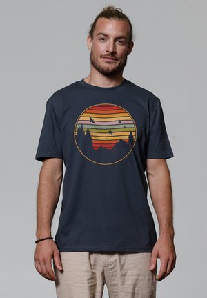 Marineblauwe katoenen t-shirt met een cirkelvormige afbeelding van een zonsondergang, vergezeld van horizontale strepen in oranje, geel, groen en silhouetten van bomen.