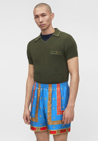 Άνδρας που φοράει πλεκτό πόλο με κοντά μανίκια σε ελιά πράσινο χρώμα με τσέπη στο στήθος και φρέσκια μπλε κοντή βερμούδα με σχέδια αλυσίδων σε χρυσό και κόκκινο χρώμα.
