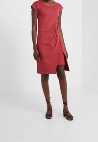 Vestido rojo ajustado con mangas cortas tipo capa, presenta un detalle retorcido en la cintura y una abertura lateral. Tejido liso y diseño hasta la rodilla.