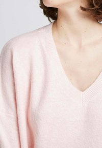 Pull v-neck rose clair avec une texture douce. Le vêtement présente une coupe décontractée et un bord côtelé le long du col et des manches.