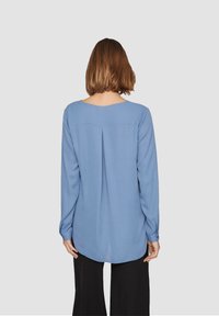 Blusa azzurra a maniche lunghe realizzata in tessuto liscio, con scollatura rotonda, dettaglio di piega sul retro e orlo arrotondato. Abbinata a pantaloni neri.