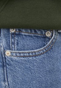 Blå denimjeans med en liten framficka, metallbeslag, synlig sömnad och en grön tygdel som delvis överlappar midjebandet.