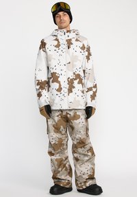 Ski-jack en -broek in het wit met een bruin camouflagedessin. De jack heeft een ritssluiting aan de voorkant en een verstelbare capuchon; de broek heeft cargopockets en een ontspannen pasvorm.