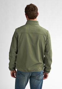 Giacca verde oliva con colletto, realizzata in tessuto liscio. Caratterizzata da dettagli di cucitura e senza hardware visibile. Indossata con jeans blu scuro.