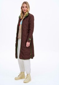 Brązowy pikowany długi płaszcz z zielonymi lamówkami, bocznymi guzikami i kołnierzem. Nosi się go na beżowym swetrze i białych spodniach, w połączeniu z beżowymi botkami.