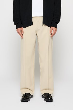 Pantalones de pierna ancha beige con costuras verticales, combinados con un cárdigan negro texturizado con cremallera al frente y zapatos negros brillantes.