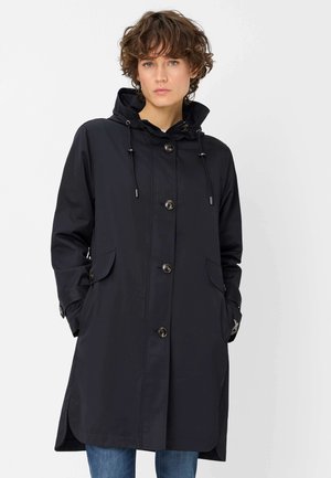 Frau trägt einen langen schwarzen Mantel mit Knöpfen, Kapuze und seitlichen Taschen, steht mit den Händen in den Taschen vor einem schlichten Hintergrund.