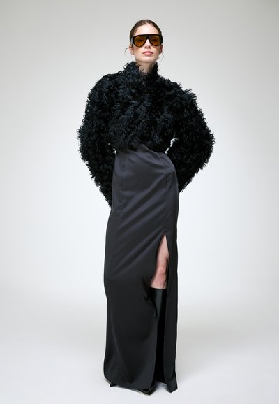 Femme portant de grandes lunettes de soleil ambrées, un haut noir texturé à manches longues et une longue jupe noire avec une fente haute, debout devant un fond uni.
