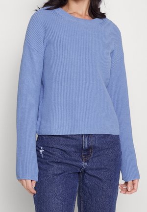 Femme portant un pull en tricot bleu clair à manches longues associé à un jean taille haute bleu foncé avec une petite déchirure sur la cuisse gauche.