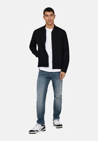 Svart jakke med glidelås og bomberstil over en hvit T-skjorte, kombinert med lyseblå jeans og svart-hvite sneakers. Minimalistisk design og passform.