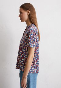 Kurzärmliges Oberteil aus blauem Stoff mit einem floralen Muster in Rot, Weiß und Gelb. Henley-Ausschnitt mit subtilen Knöpfen und einer lockeren Passform.