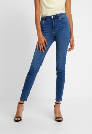 Kobieta w niebieskich dopasowanych spodniach jeans z średnim stanem i czarnych szpilkach z paskiem wokół kostki, stojąca na tle jednolitego jasnego tła.