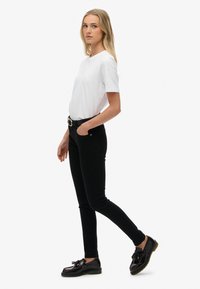 Camiseta blanca de manga corta, jeans ajustados negros con cinturón y zapatos negros brillantes con detalle de borlas, sobre un fondo blanco liso.