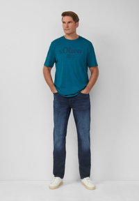 Türkises T-Shirt mit kurzen Ärmeln und schwarzem Text, kombiniert mit dunkelblauen Jeans und weißen Sneakers. Das Design ist lässig und schlicht.