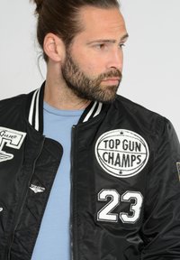 Zwarte bomberjack met witte accenten, voorzien van een "TOP GUN CHAMPS" patch, het nummer "23" en een ritssluiting. Draagt over een lichtblauw t-shirt.