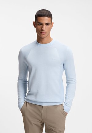 Jonge man die een lichtblauwe trui met lange mouwen en beige broek draagt, staand met handen in de zakken tegen een effen achtergrond.