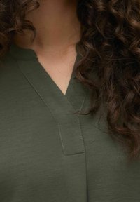 Haut vert olive avec un design en V, présentant des détails de couture subtils et une texture lisse. Cheveux bouclés partiellement visibles.