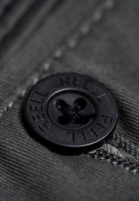 Schwarzer Kunststoffknopf mit dem Aufdruck "REELL", der ein Vierlochdesign zeigt, auf einem strukturierten, dunkelgrauen Stoffhintergrund angenäht.