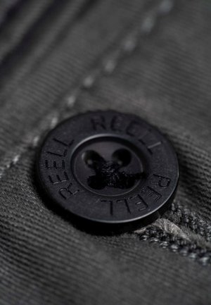Botón de plástico negro con la inscripción "REELL" en relieve, que presenta un diseño de cuatro agujeros, cosido sobre un fondo de tela texturizada de color gris oscuro.