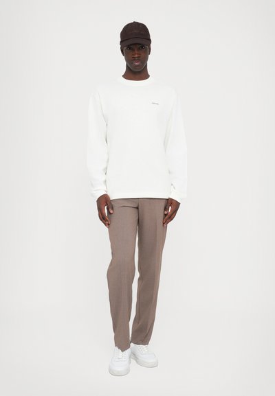 White long-sleeve shirt, brown cap, brown patterned trousers, and white sneakers. Casual fit with minimal branding on the shirt.
