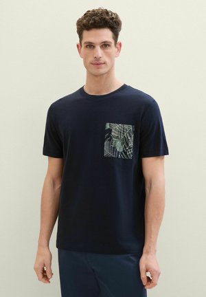 Hombre con pelo rizado que lleva una camisa de manga corta azul marino con un bolsillo en el pecho estampado, de pie contra un fondo claro y liso.