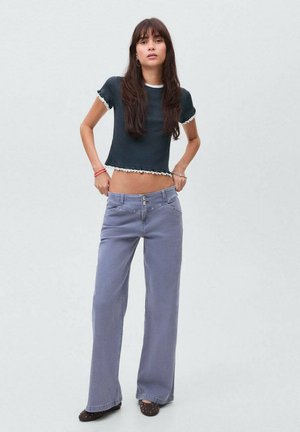 Femme aux longs cheveux noirs portant un t-shirt noir court et un jean bleu oversize taille haute, se tenant devant un fond uni.