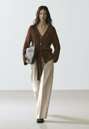 Donna che cammina indossando un cardigan marrone lavorato a maglia, pantaloni beige a gamba larga, scarpe nere e tenendo una grande borsa clutch nera con texture.
