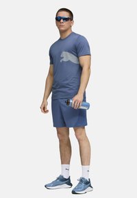 Herre aktivtøj outfit bestående af en blå kortærmet skjorte med et lyseblåt grafisk motiv, blå shorts og blå sneakers. Vandflaske i hånden.