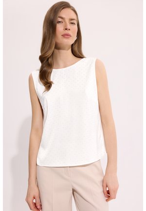 Kobieta o długich falowanych włosach nosi białą bezrękawnikową bluzkę z subtelnym wzorem kropek oraz beżowe spodnie na jasnym, jednolitym tle.