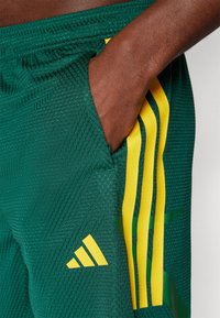 Zeleni športni kratki hlače iz teksturirane mrežaste tkanine z rumenimi tremi črtami in majhnim adidas logotipom na žepu na levi strani.
