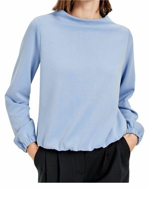 Kobieta w jasnoniebieskiej bluzce z długim rękawem z elastycznymi mankietami i wykończeniem na dole, w połączeniu z czarnymi, wysokim stanem spodniami, z rękami w kieszeniach.