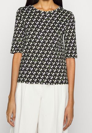 Frau trägt ein schwarzes Kurzarmshirt mit weißen geometrischen Sternmustern und kleinem grünen "SPORTS"-Schriftzug, kombiniert mit weißen Hosen.