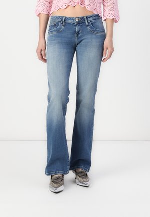 Kobieta nosząca jasnoniebieskie wyblakłe jeansy typu bootcut, różowy koronkowy krótki top oraz srebrne metaliczne, spiczaste trampki.