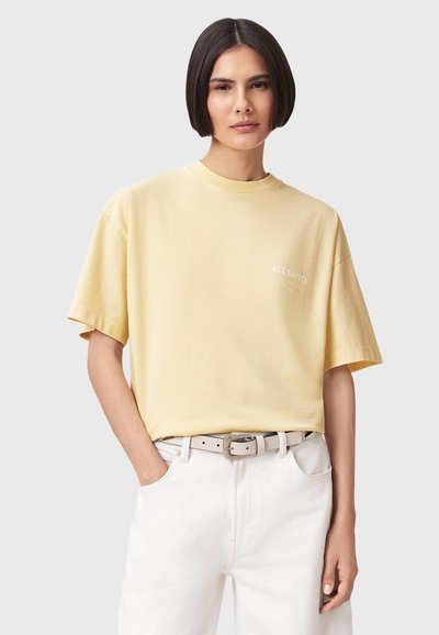Jeune personne aux cheveux courts et foncés portant un t-shirt oversize jaune pâle rentré dans un pantalon blanc taille haute avec une ceinture beige.