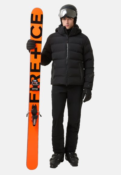 Eine Person in einer schwarzen Daunenjacke hält einen orangefarbenen Ski mit schwarzer Schrift. Sie trägt schwarze Skihose, Handschuhe und einen Helm mit einer Ski-Brille.