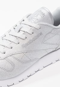 Silver athletic shoe with a textured finish, featuring white laces and branding. Soft upper material and a thick white rubber sole.